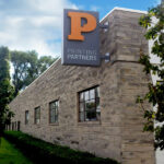Printing Partners building at 929 W 16th Street, Indianapolis, IN, showcasing the prominent 'P' logo on a stone exterior with lush greenery in the foreground.
