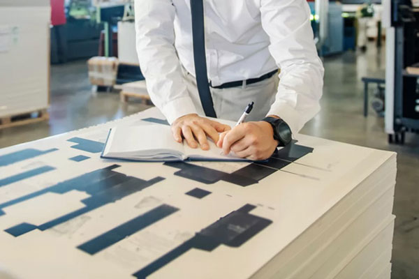 Individual in a white shirt and tie writing in a notebook on top of stacked paper sheets.