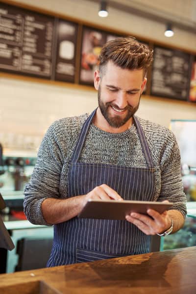 A man wearing an apron smiles while using a tablet in a restaurant setting.
